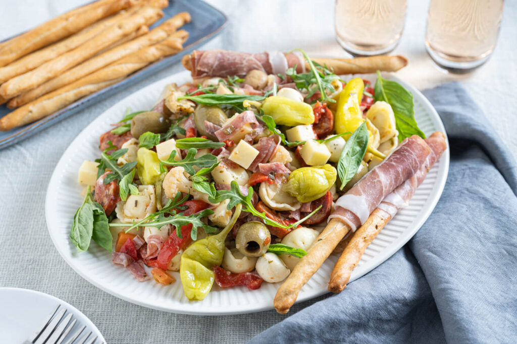 Plated antipasto salad with assorted meats, cheeses, olives, pickled items, arugula, and prosciutto-wrapped breadsticks