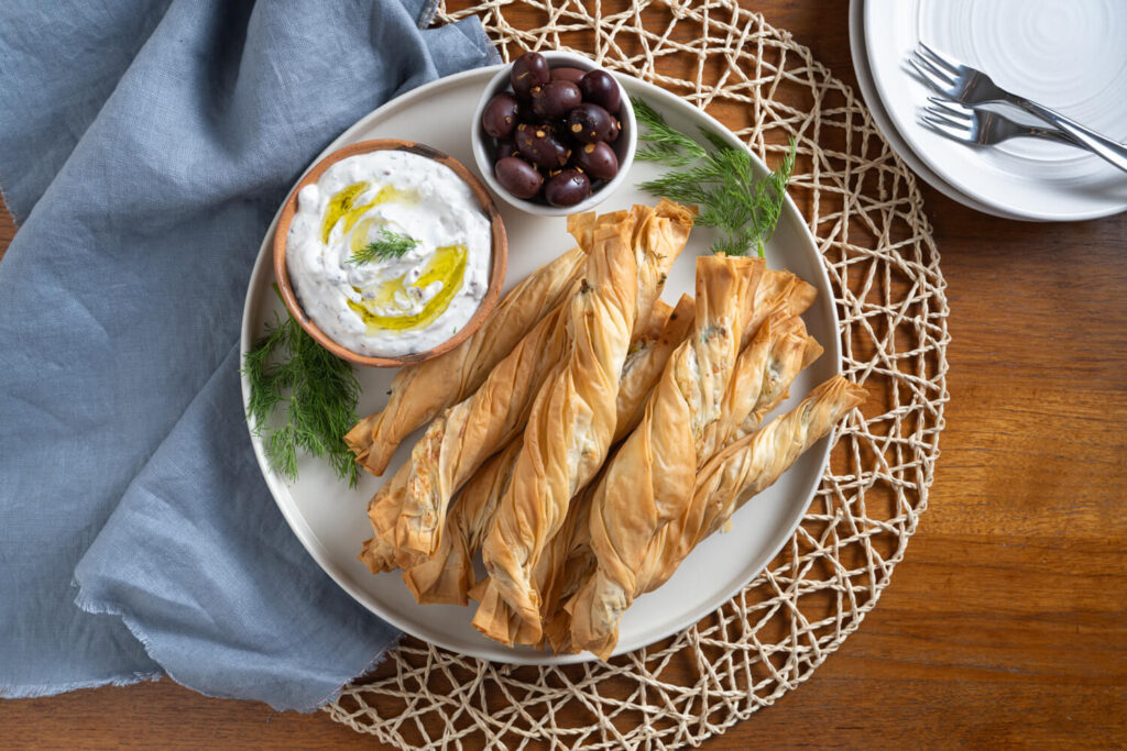 Overhead image of a plate of phyllo pastry twists with a small bowl filled with black olives and a small bowl of tzatziki dip for a potluck appetizer
