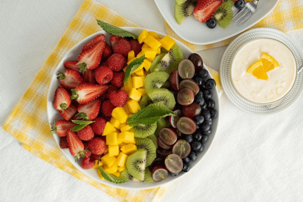 Summer Recipe: Overhead image of rainbow fruit salad with fresh berries, kiwi, mango, and grapes, with a small bowl of honey yogurt sauce.