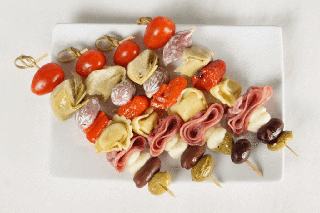 Overhead view of four skewers on a plate stacked with cherry tomatoes, tortellini, olives, bocconcini, and various meats