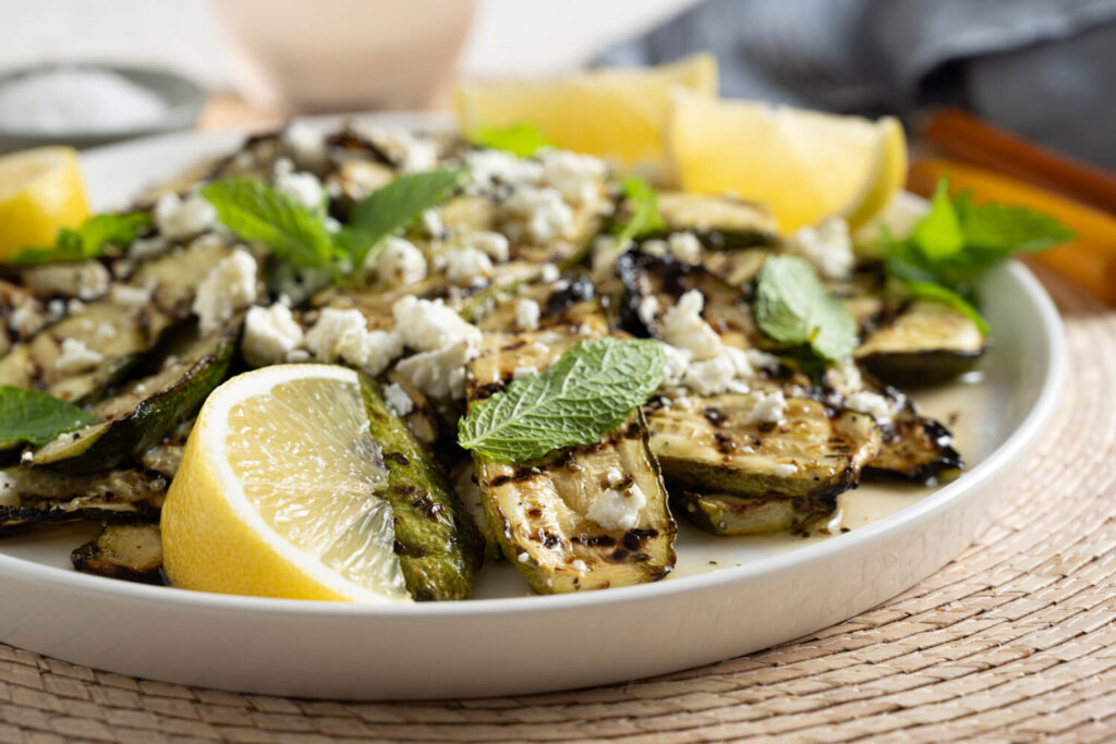 Close up of grilled zucchini salad garnished with mint leaves and feta cheese and served with wedges of lemon.