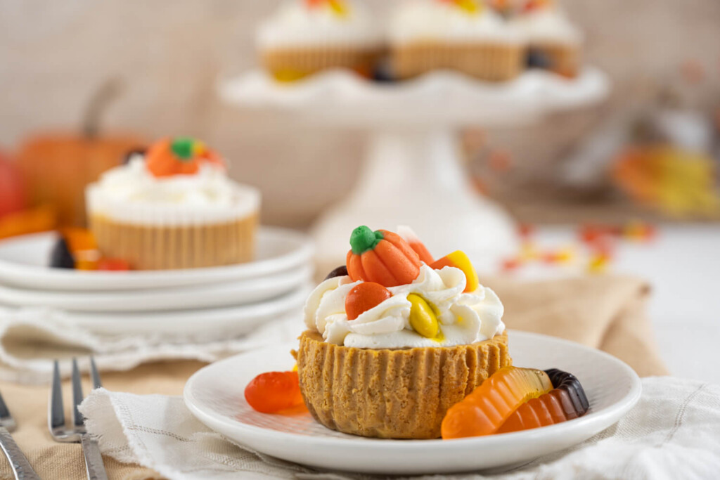 Mini pumpkin spice cheesecake on a plate decorated with whipped cream, Halloween gummy worm, and mini candy pumpkin.