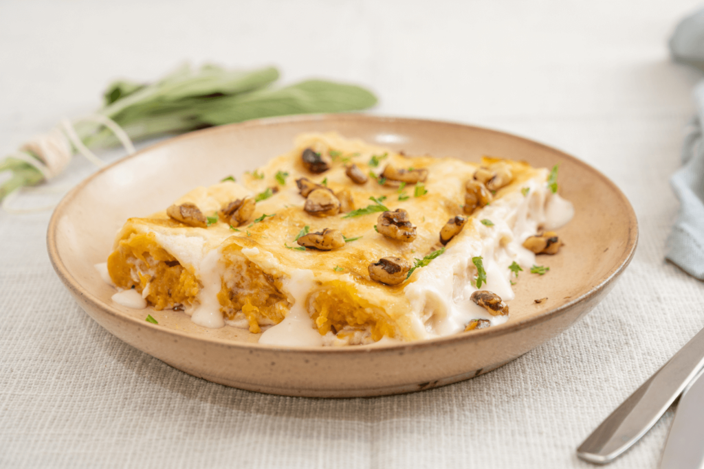 Tan stoneware dish with three rolls of butternut squash cannelloni made with a creamy Alfredo sauce and garnished with toasted walnuts.