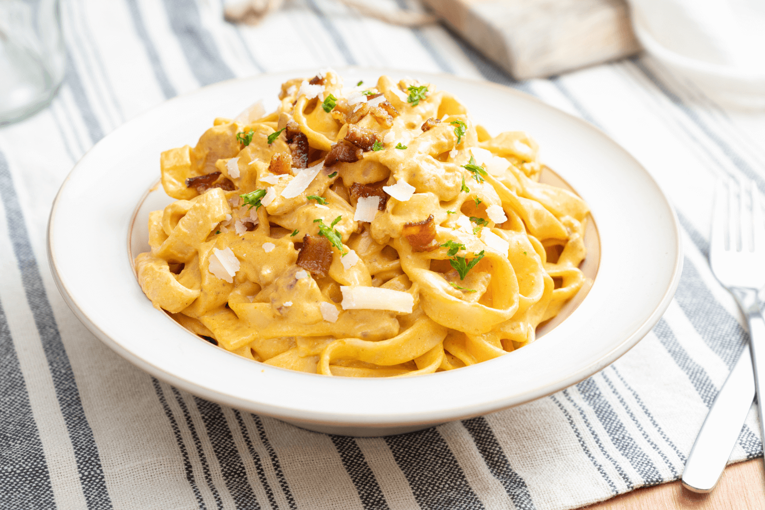 White dish of creamy pumpkin chicken tagliatelle garnished with bacon bits and Parmesan cheese on a striped placemat.