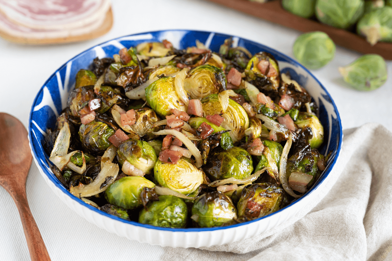 The image shows a blue and white bowl filled with roasted Brussels sprouts cooked with crispy pancetta and onions. Beside the bowl is a wooden spoon.