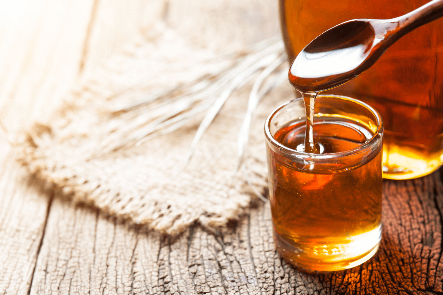 Wooden spoon dipping into small glass of maple syrup on wooden table.