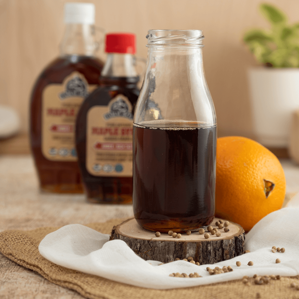 Glass bottle of maple syrup on wooden coaster with coriander seeds and whole navel orange.