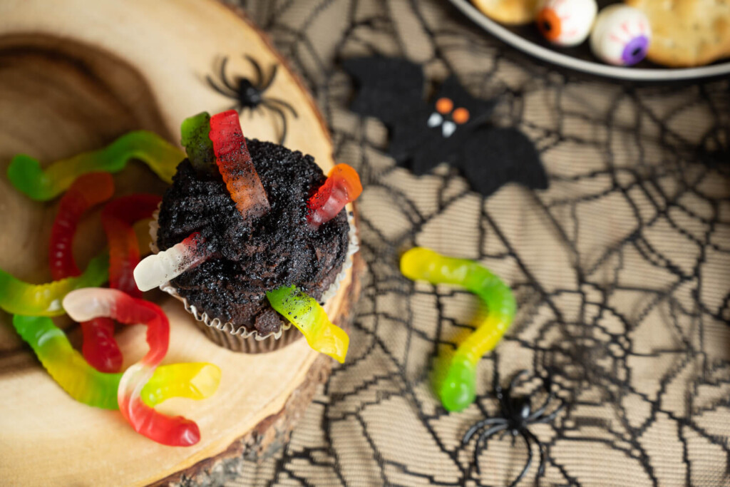 Overhead image of a chocolate cupcake with gummy worms surrounded by Halloween decor.