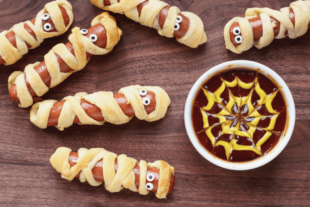 Fun food for kids. Mummy hot dogs lying on a rustic table with a bowl of ketchup and mustard dip, with spider web design. Top view, flay lay position.