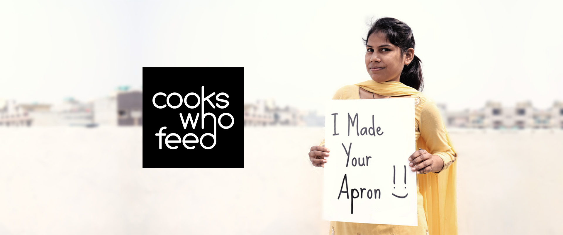 Cooks who Feed. Woman who is part of the cooks who feed community is holding a sign that reads "I made your apron".