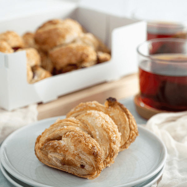 The image shows a stck of two pale blue plated in the foreground upon which is a sugar-crusted strudel. In the background out of focus is a clear cup of tea and an open box containing more strudels.