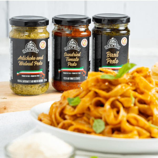 selection of jarred Farm Boy Pestos in focus in background behind plate of cooked pasta with pesto.