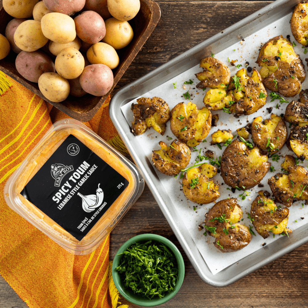 Overhead image of baking sheet with smashed baby potatoes garnished with fresh herbs. Nearby, a container of Farm Boy Spicy Toum