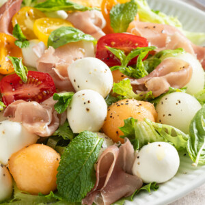 A colourful and vibrant salad arranged on a white plate. The salad features a variety of melon balls, including canary melon, cantaloupe, and honeydew, interspersed with small bocconcini cheese balls. Thin slices of savory prosciutto are delicately draped over the melon and cheese. The salad is garnished with fresh basil leaves and mint leaves with a drizzle of the Farm Boy Prosecco Vinegar, creating a visually appealing and appetizing dish.