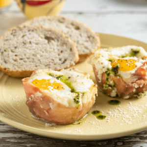 Two cheesy prosciutto cups with two slices of bread on a pale yellow plate. The cheesy prosciutto cups have eggs, vibrant fresh pesto, and grated Parmigiano Reggiano cheese. This is an easy breakfast dish that is satisfying and savoury.