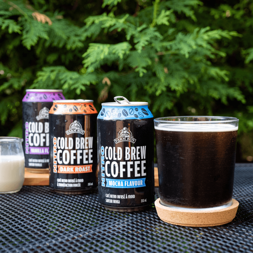 Three cans of Farm Boy Nitro Brew Coffee on a table outside with a short glass filled with cold brew coffee