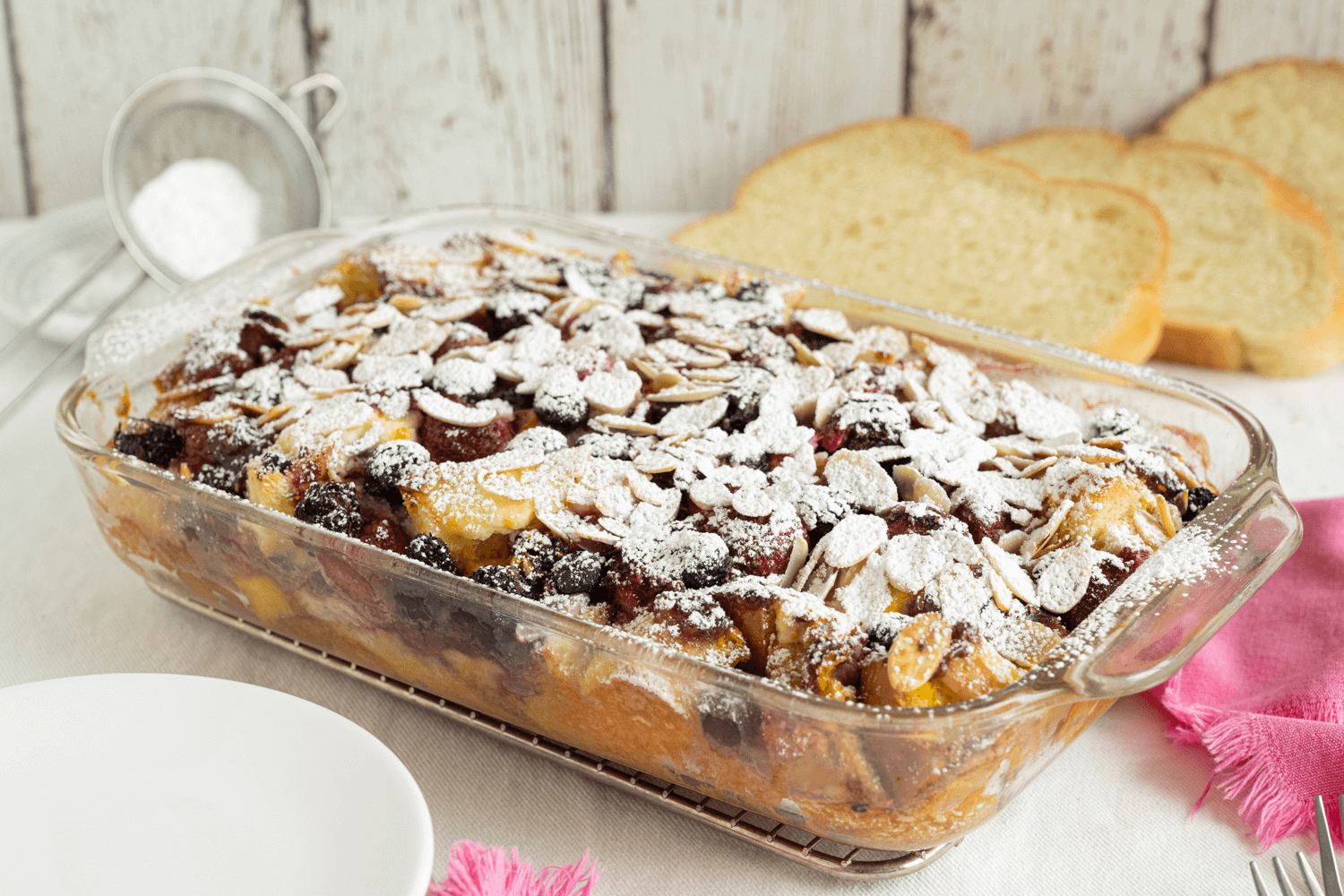 mixed berry strata dessert. The image shows a glass casserole dish filled with mixed berry strata, dusted with icing sugar. In the background are slices of fresh challah bread and a pink scarf.