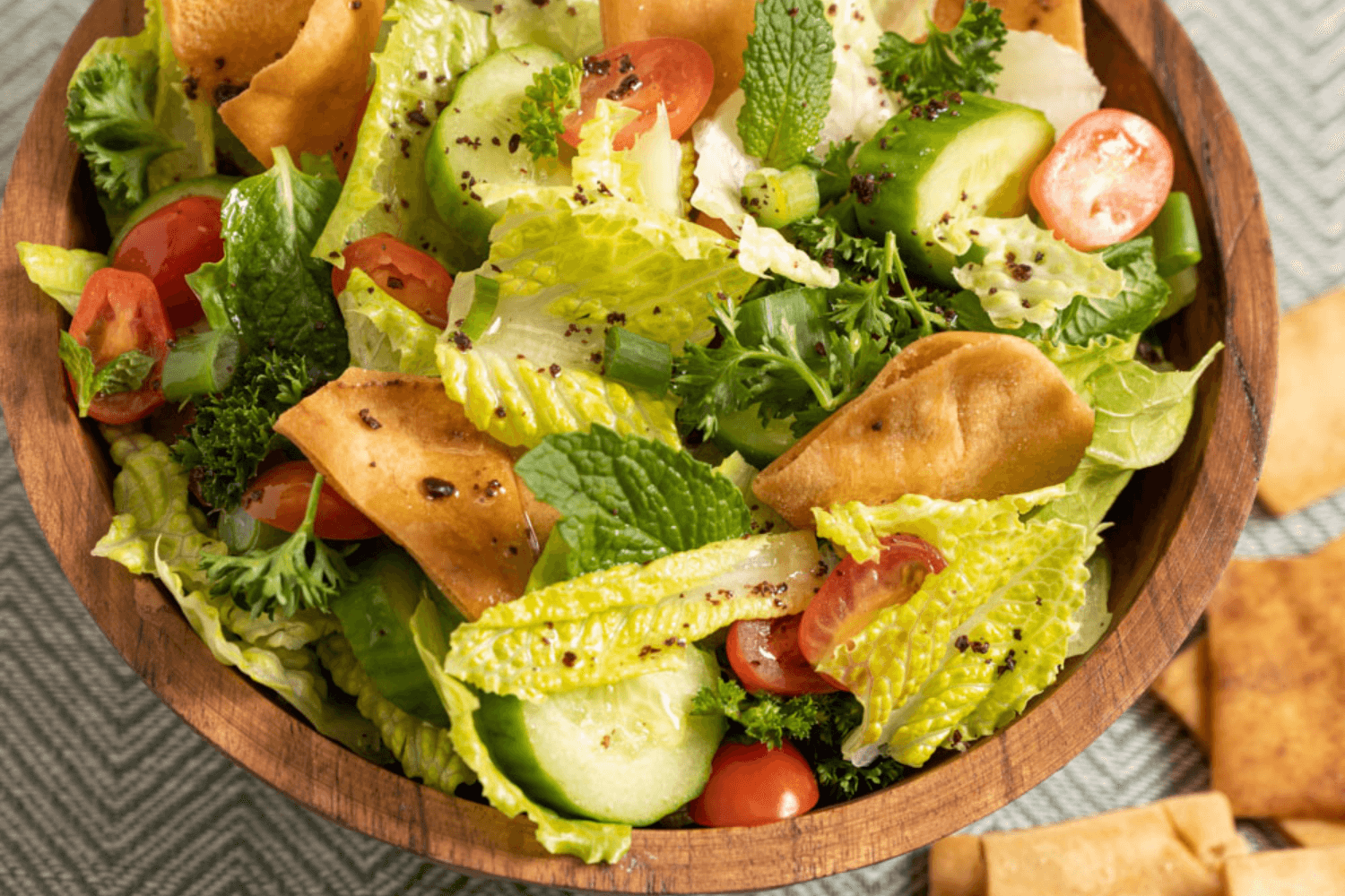 The image shows a closeup of a wooden bowl filled with chopped lettuce, cucumber coins, sliced cherry tomatoes, and fattoush chips.