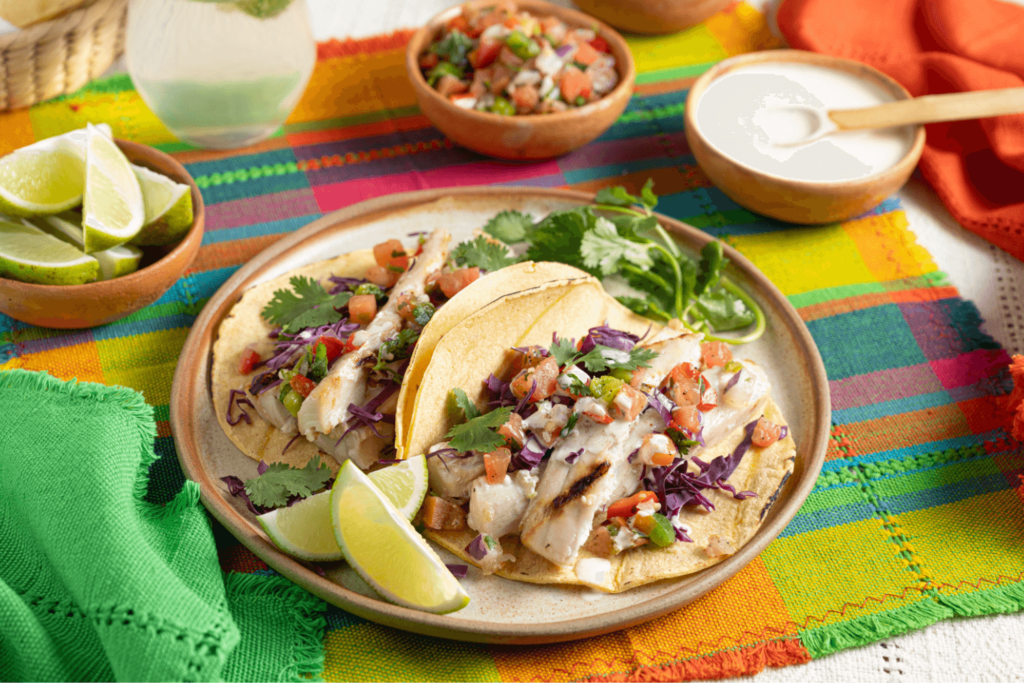The image shows a stoneware plate with two fish tacos topped with fresh pico de gallo and a wedge of lime. The plate is on a colourful woven placemat.