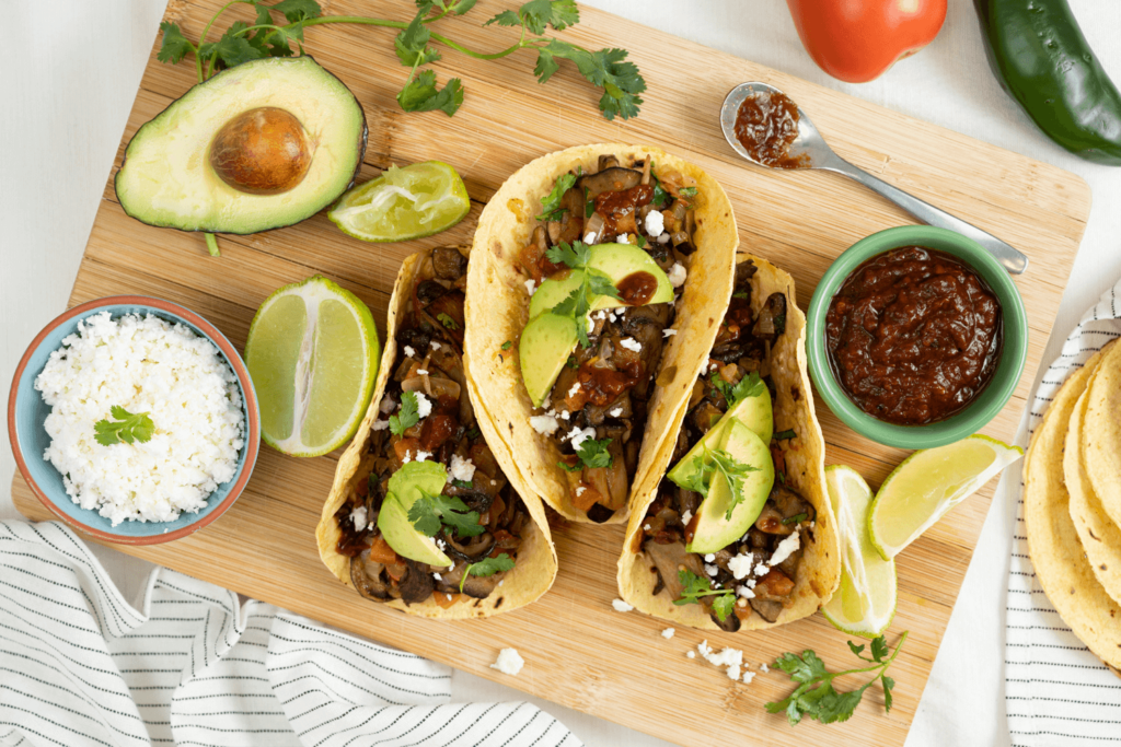 The image is shot from above and shows three mushroom tacos on a wooden cutting board. Also on the cutting board is half an avocado, a small bowl of feta cheese, a small bowl of salsa, and fresh wedges of lime.
