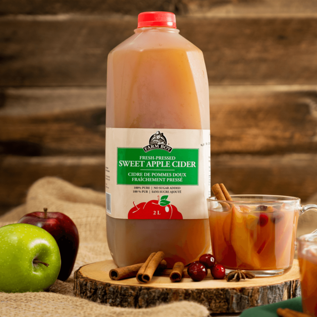 Jug of Farm Boy Sweet Apple Cider on cross-section of wood beside a clear glass mug of mulled cider with apple slices, cranberries, and cinnamon sticks.