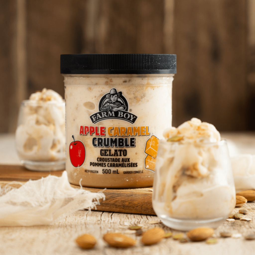 Container of Farm Boy Apple Caramel Crumble Gelato on wooden table with wooden background. In foreground to the right and slightly out of focus is a small glass with the gelato. Scattered almonds and pumpkin seeds are in the foreground.