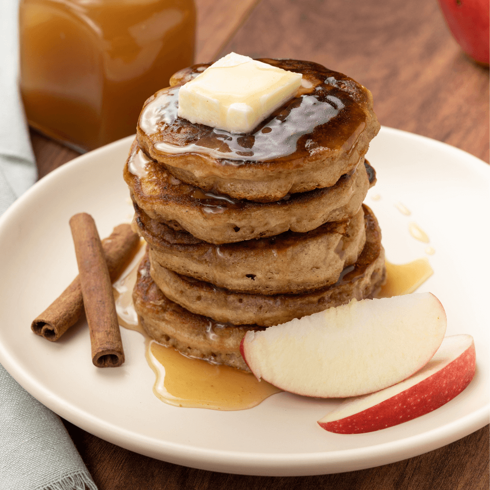 The image shows a white plate with a stack of five apple pancakes. There is a pat of butter on top of the pancakes and an apple cider sauce. Beside the pancakes are two wedges of apple and two crossed cinnamon sticks.