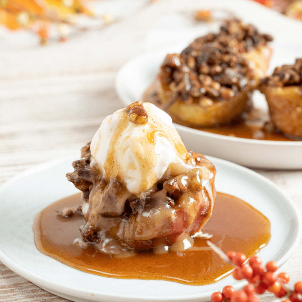 The image shows a plate with a baked apple sitting in a pol of caramel sauce. The apple is topped with nuts, a scoop of vanilla ice cream, and caramel sauce.