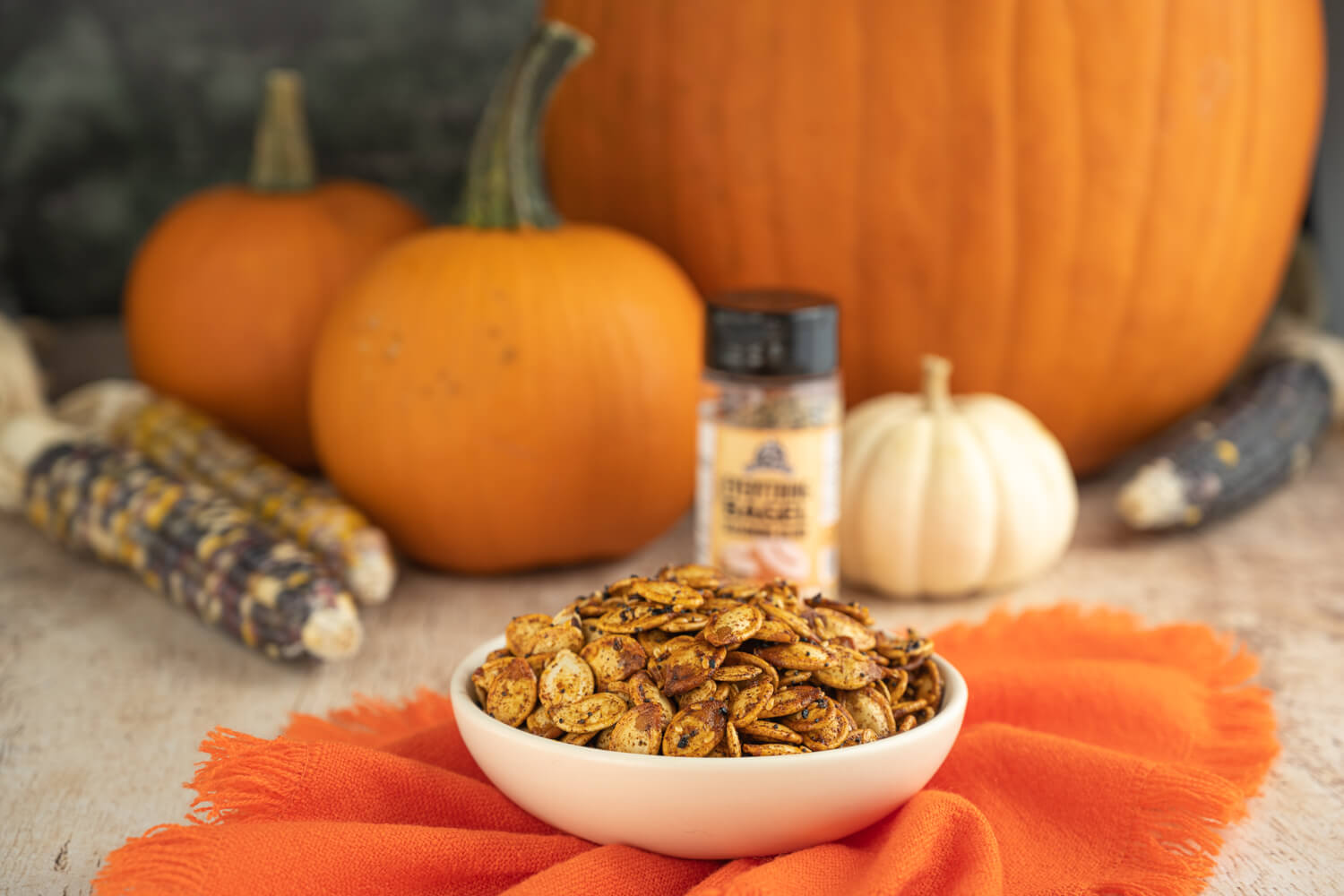 Bowl of roasted pumpkin seeds on an orange cloth with various pumpkins in the background.
