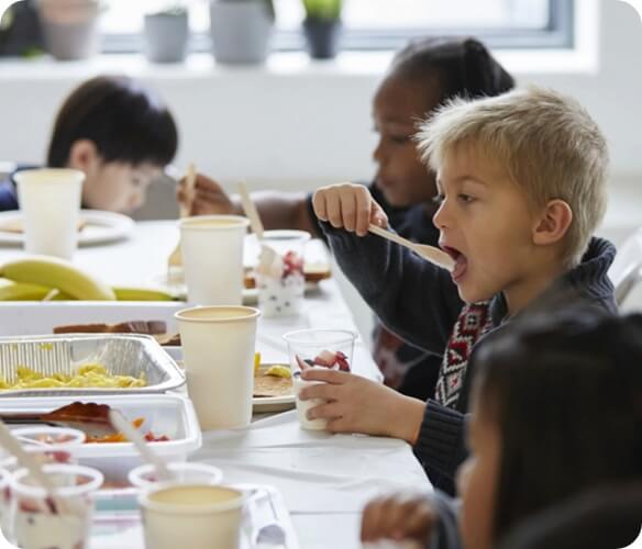 Image of kids sitting at a table eating their lunch from containers and cups.