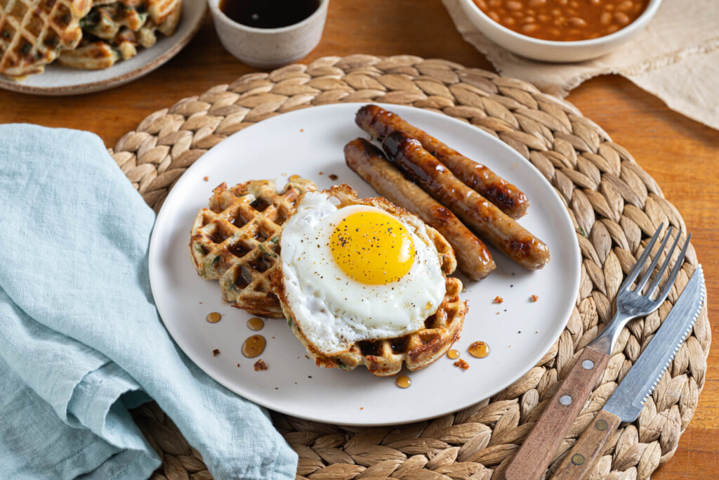 Wicker placemet with white plate with two savoury spinach and cheese waffles with fried egg and sausage links for yummy brunch