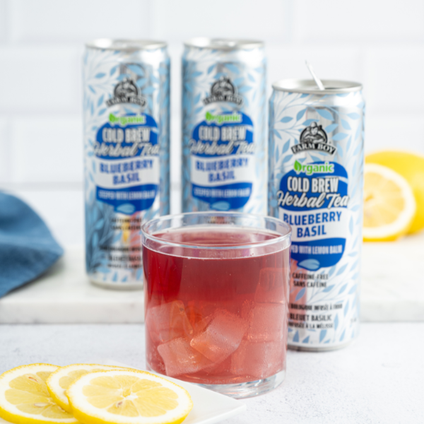 Cans of Farm Boy Blueberry Basil tea behind short glass filled with purple-red iced tea