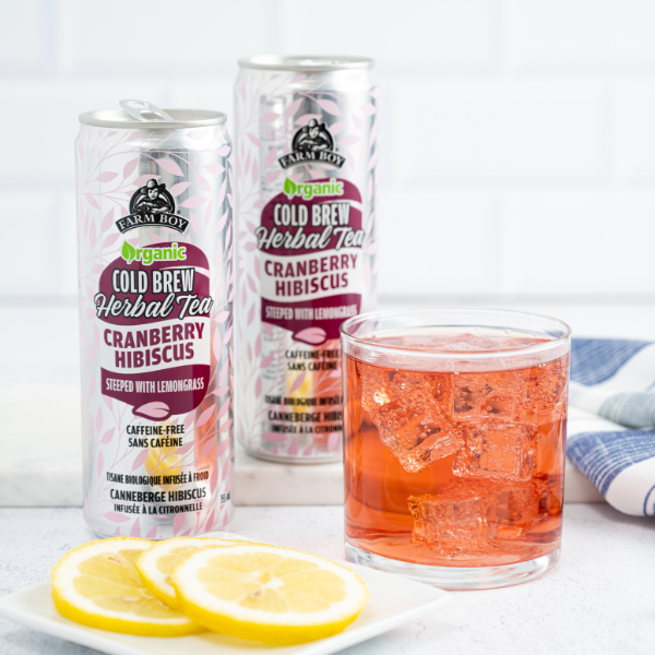 Cans of Farm Boy Cranberry Hibiscus tea beside short glass of orange-red iced tea