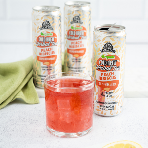 Cans of Farm Boy Peach Hibiscus Tea behind short glass filled with rosy-coloured iced tea