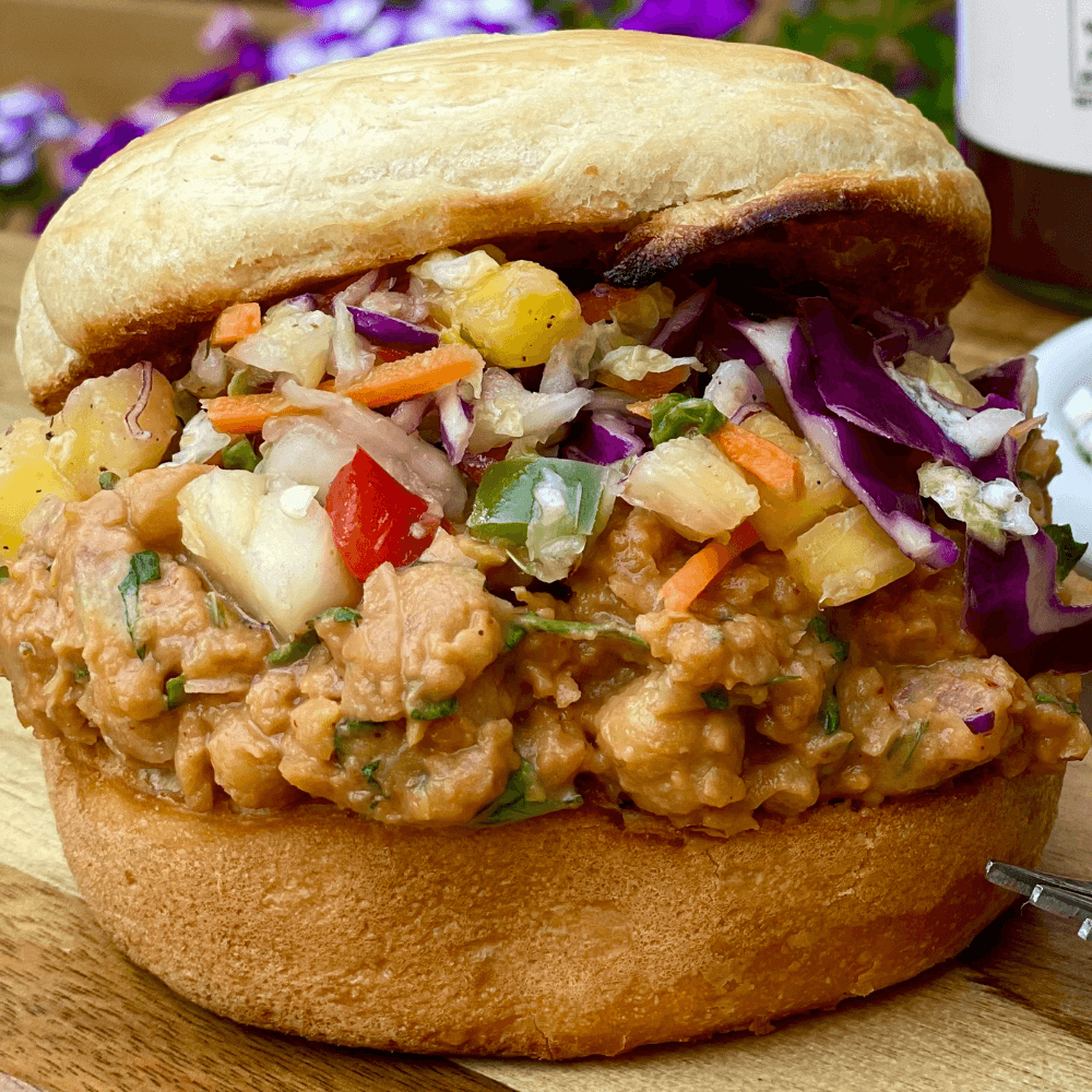 The image shows a closeup of a toasted burger bun with a chickpea mixture and colourful slaw. The sandwich is on wooden table.