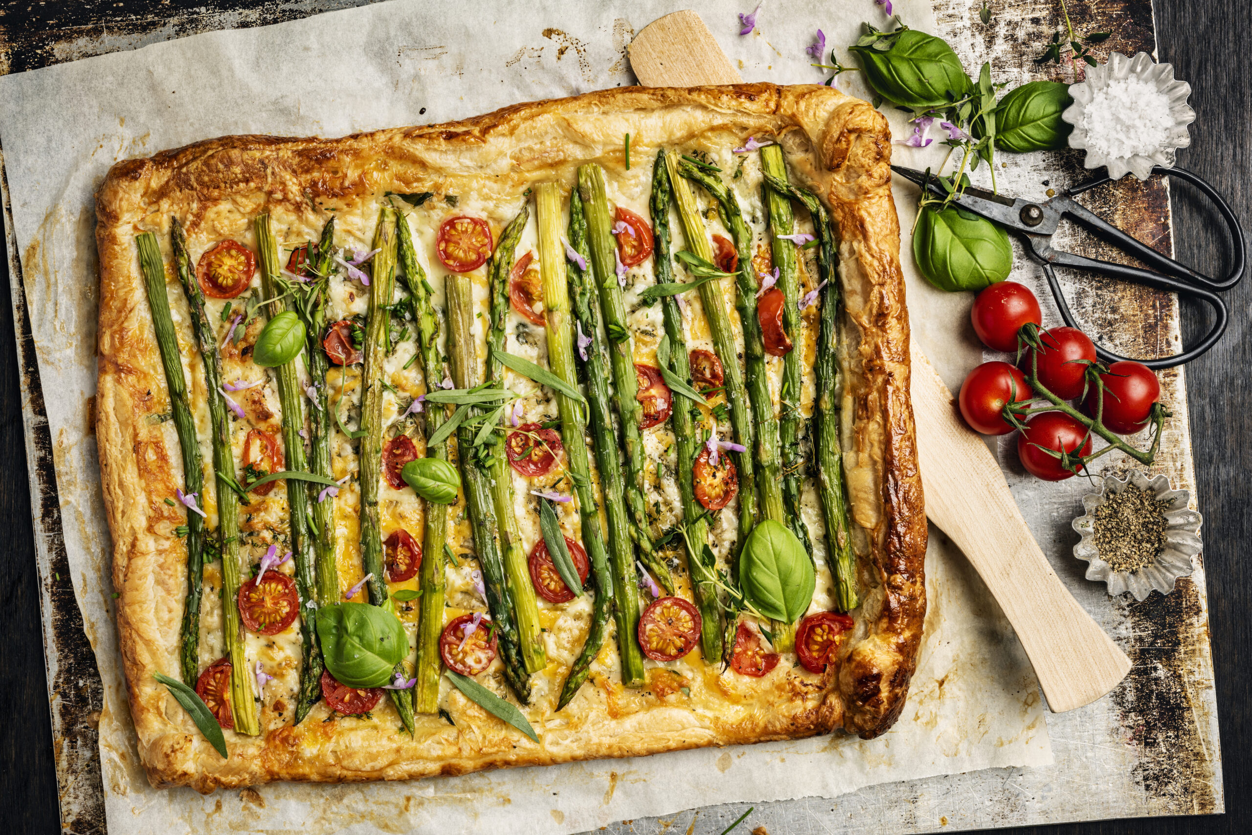 Overhead view of an asparagus, cheese and tomato tart. A shop bought puff pastry dough has been used as a base for the tart. Recipe; roll the  puff pastry out and make a thin cut not too deep about 2cm from the edge all around the pastry. Within the cuts prick the base of the pastry with a fork, lightly cover the pastry with some olive oil and herbs such as, chives, cress, thyme and parsley. Grate some cheese over the base such as gruyere or red cheddar and place the lightly cooked  asparagus spears that have have had the last few centimetres of woody stalk removed nose to tail on top of the cheese. Then chopped cherry tomatoes added, some shavings of fresh parmesan and some salt and pepper for seasoning. Fold over the 2 cm edges and brush some egg yolk over the pastry if there is any egg yolk left it can be poured over the asparagus. Cook for around 15 to 20 minutes at 180 degrees, delicious!