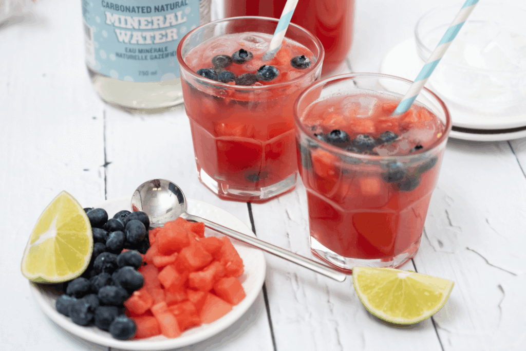 Two short glasses of sparkling watermelon drink with blueberries and blue striped straws on white wooden table