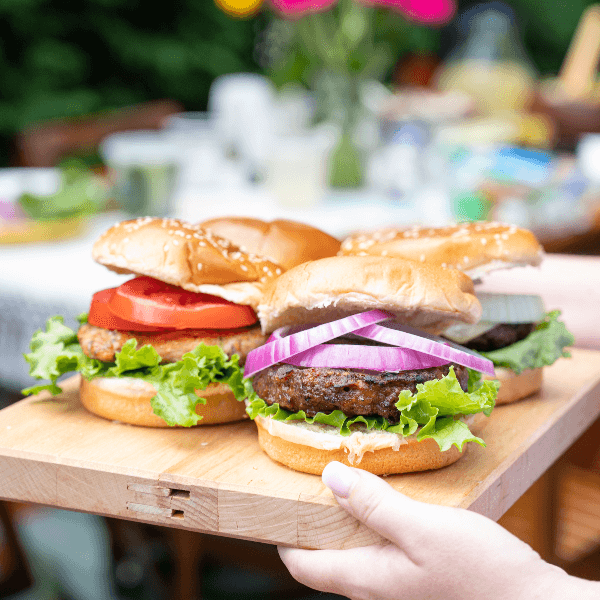 Four dressed burgers on a wooden serving board being carried by a person