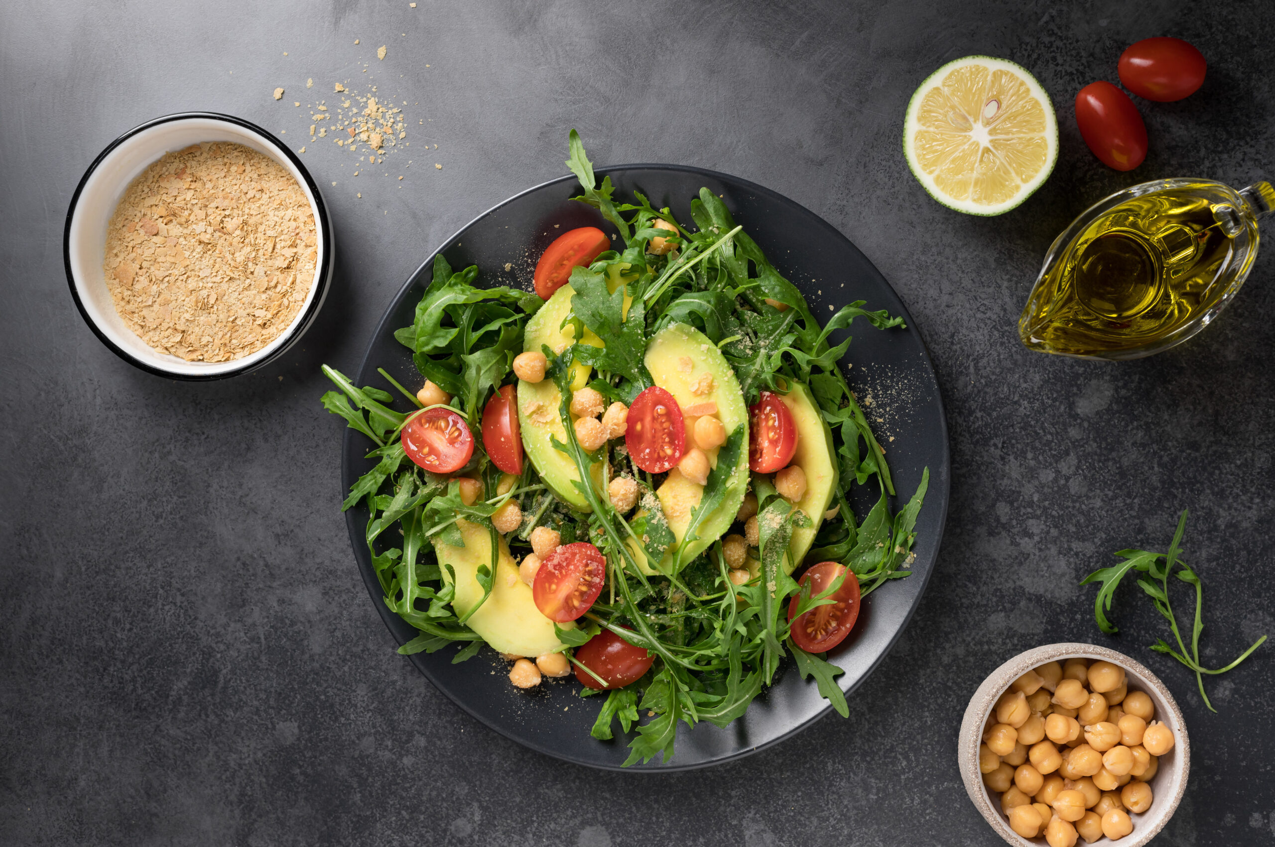 Vegan salad with avocado, arugula, tomatoes, chickpeas and nutritional yeast flake on dark background. Mediterranean cuisine. flat lay