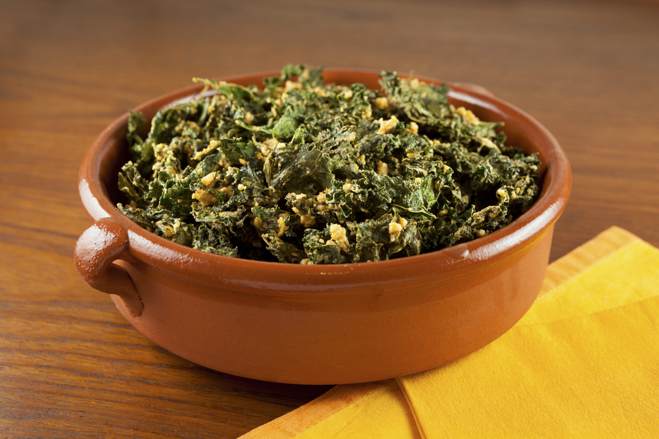 Homemade kale chips in an earthen serving bowl propped with a yellow napkin. Kale chips are made with nutritional yeast.