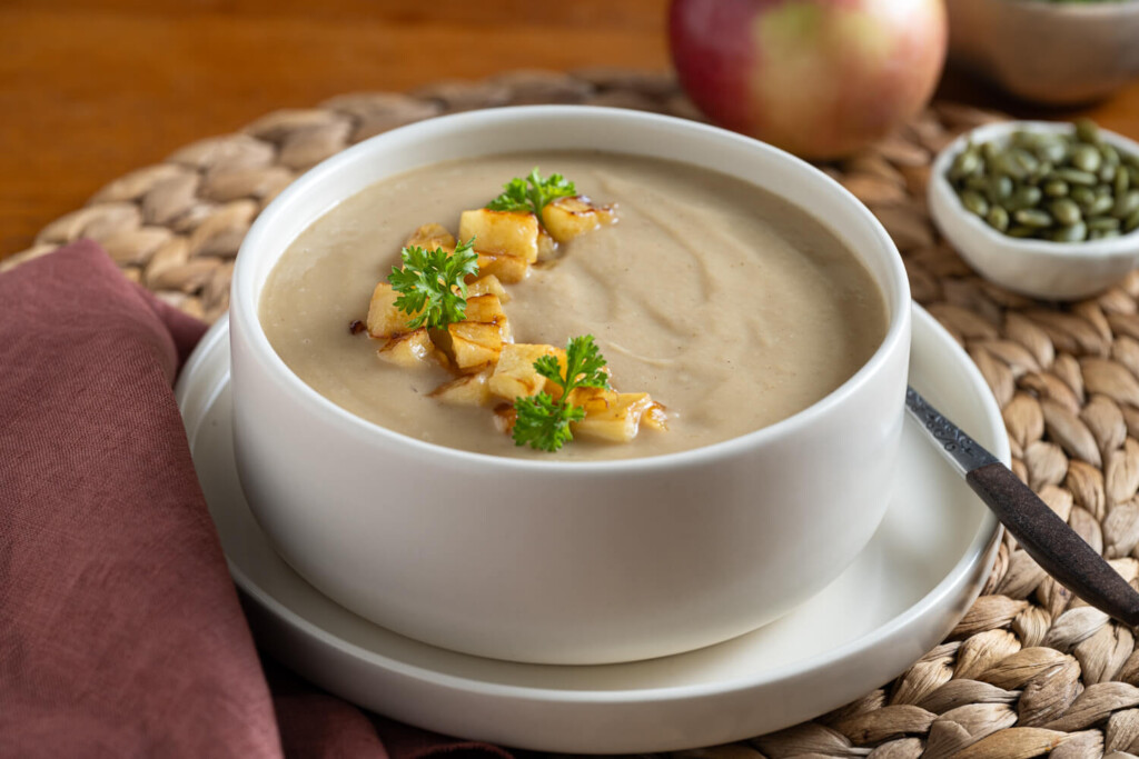 White plate with white bowl filled with beige coloured soup garnished with apples and herbs