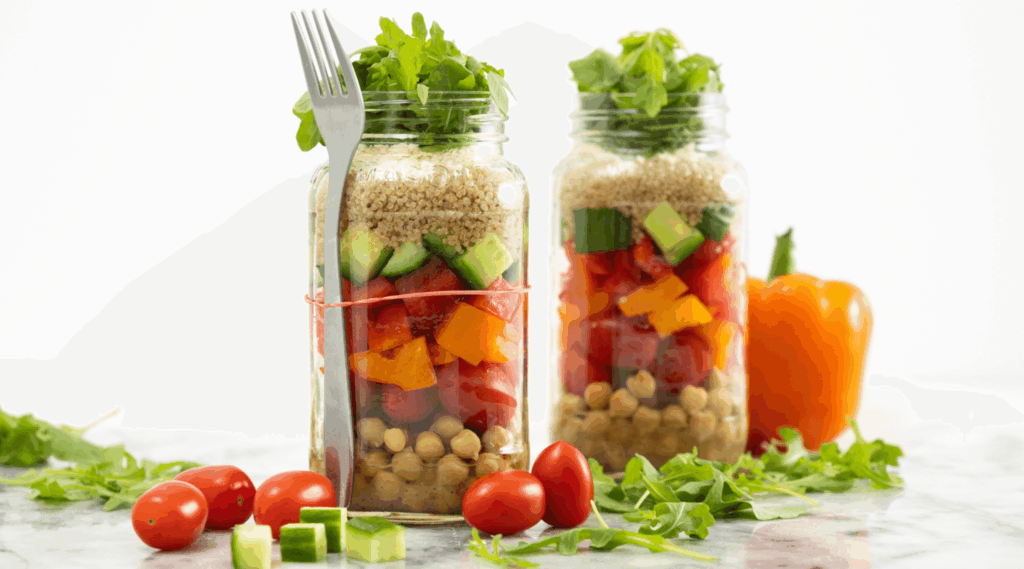 Two glass jars of layered salad with quinoa, chickpeas, bell peppers, cucumbers, tomatoes, and leafy greens