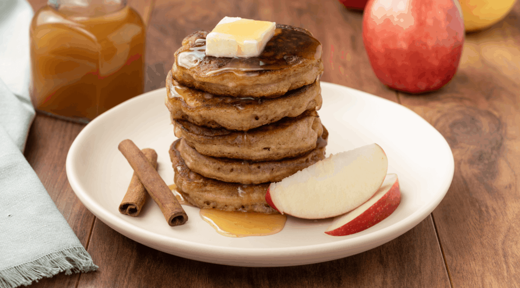 White plate with stack of apple pancakes with pat of butter on top and apple cider syrup