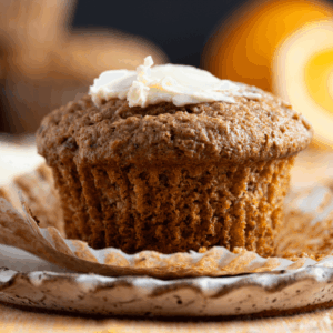 Small plate with fluted edges with an unwrapped carrot pecan bran muffin on it, topped with a pat of butter. Oranges and more muffins are visible in background
