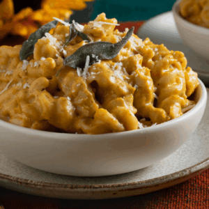 Bowl of pumpkin mac and cheese made with fussili pasta and garnished with fried sage leaves and grated Parmesan cheese with sunflowers in background