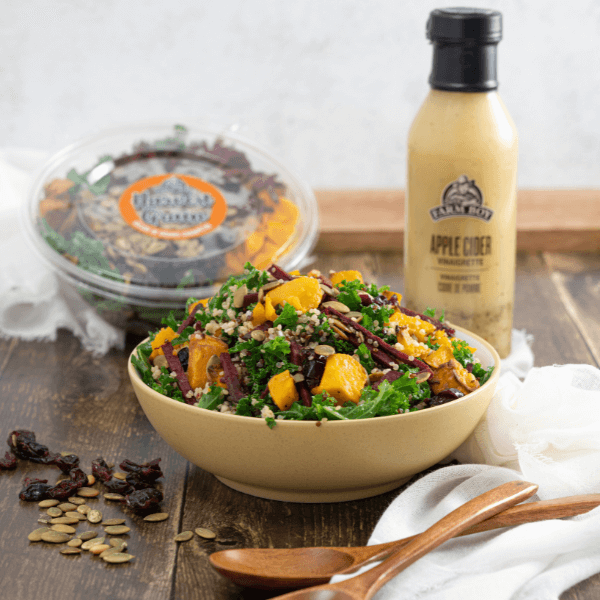 Table with bowl of salad containing butternut squash, beets, and kale with container of Farm Boy Harvest Grain Salad and bottle of Farm Boy Apple Cider Vinaigrette in background