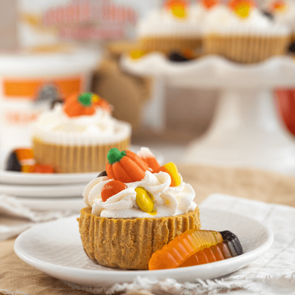 Miniature pumpki spice cheesecake with Halloween candies on a white plate with cake stand of pumpkin spice cheesecakes in background