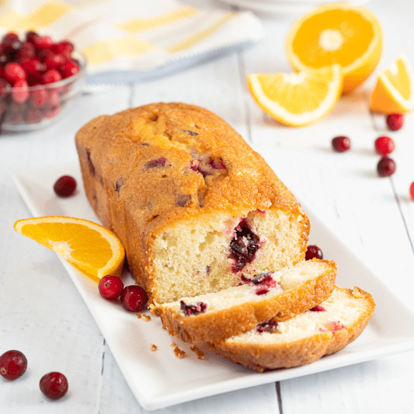 Rectangular plate with Cranberry Orange Loaf Cake with two slices cut. Surrounded by fresh cranberries and slices of orange