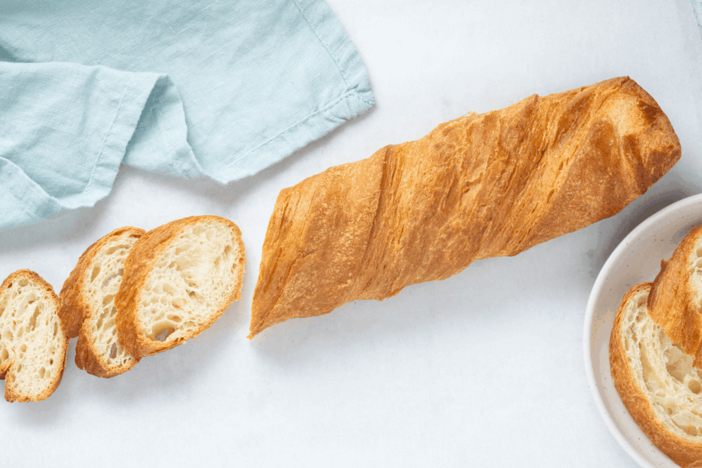 Thawed croissant demi baguette with three slices cut from it on white table with pale blue cloth