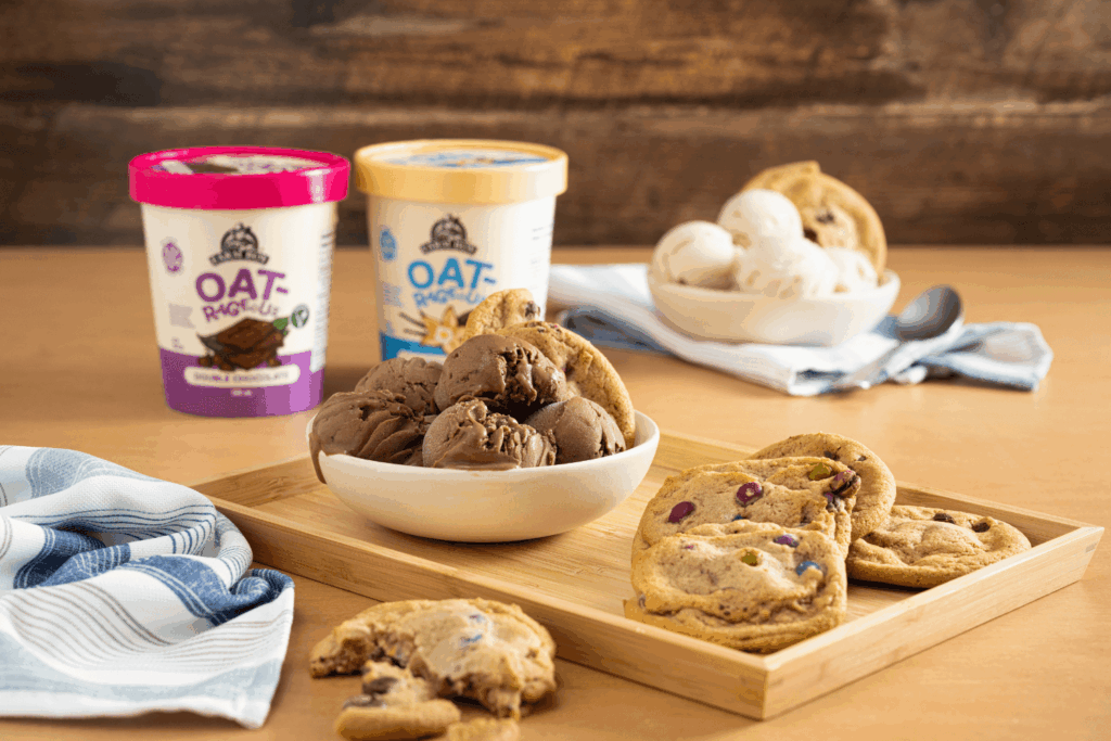 Wooden serving tray with bowl of chocolate ice cream and plant based cookies. Two pints of Farm Boy Oatrageous Frozen Dessert in background